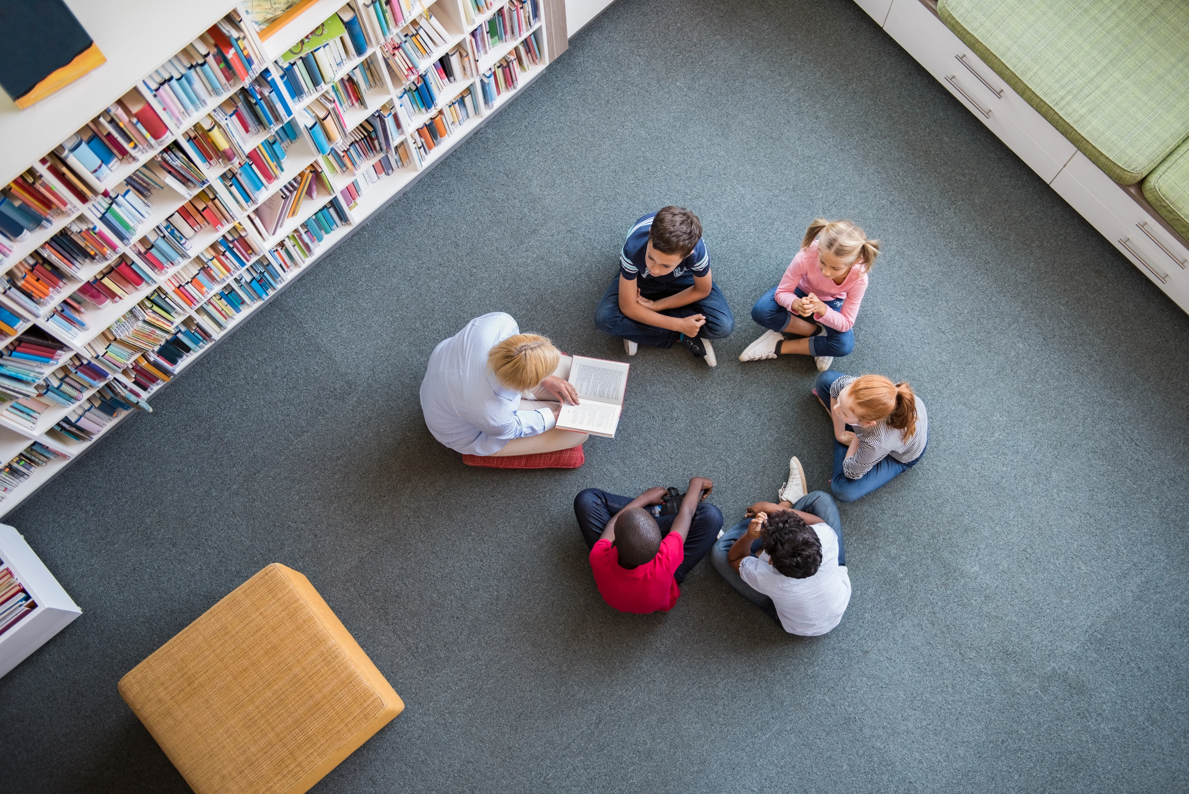 Fünf Kinder sitzen mit einer erwachsenen Person in der Schulbibliothek und bekommen ein Buch vorgelesen.
