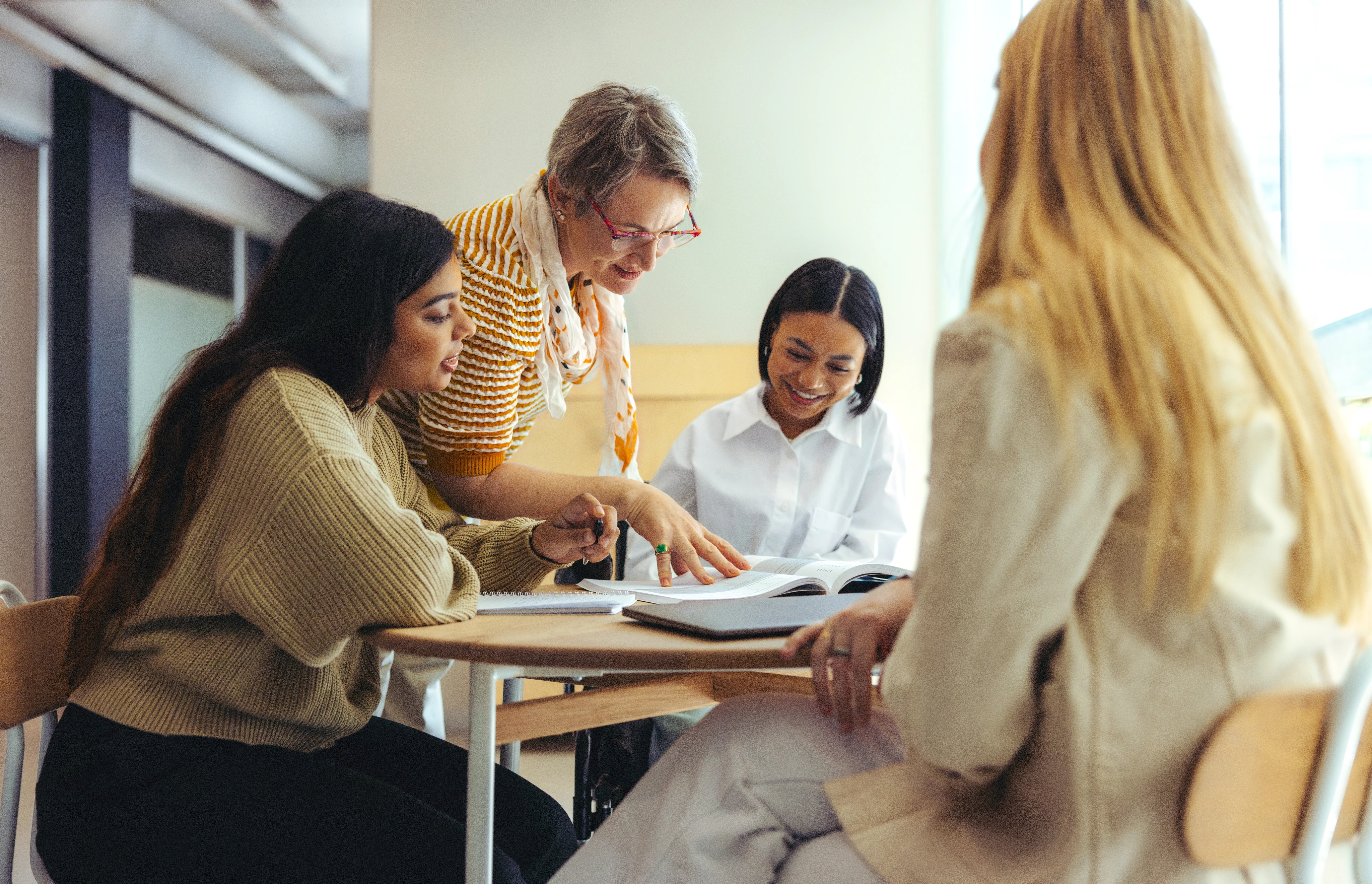 Foto einer Gruppe Schülerinnen, die an einem Tisch zusammensetzen. Am Tisch steht eine Frau in hilft bei den Aufgaben.