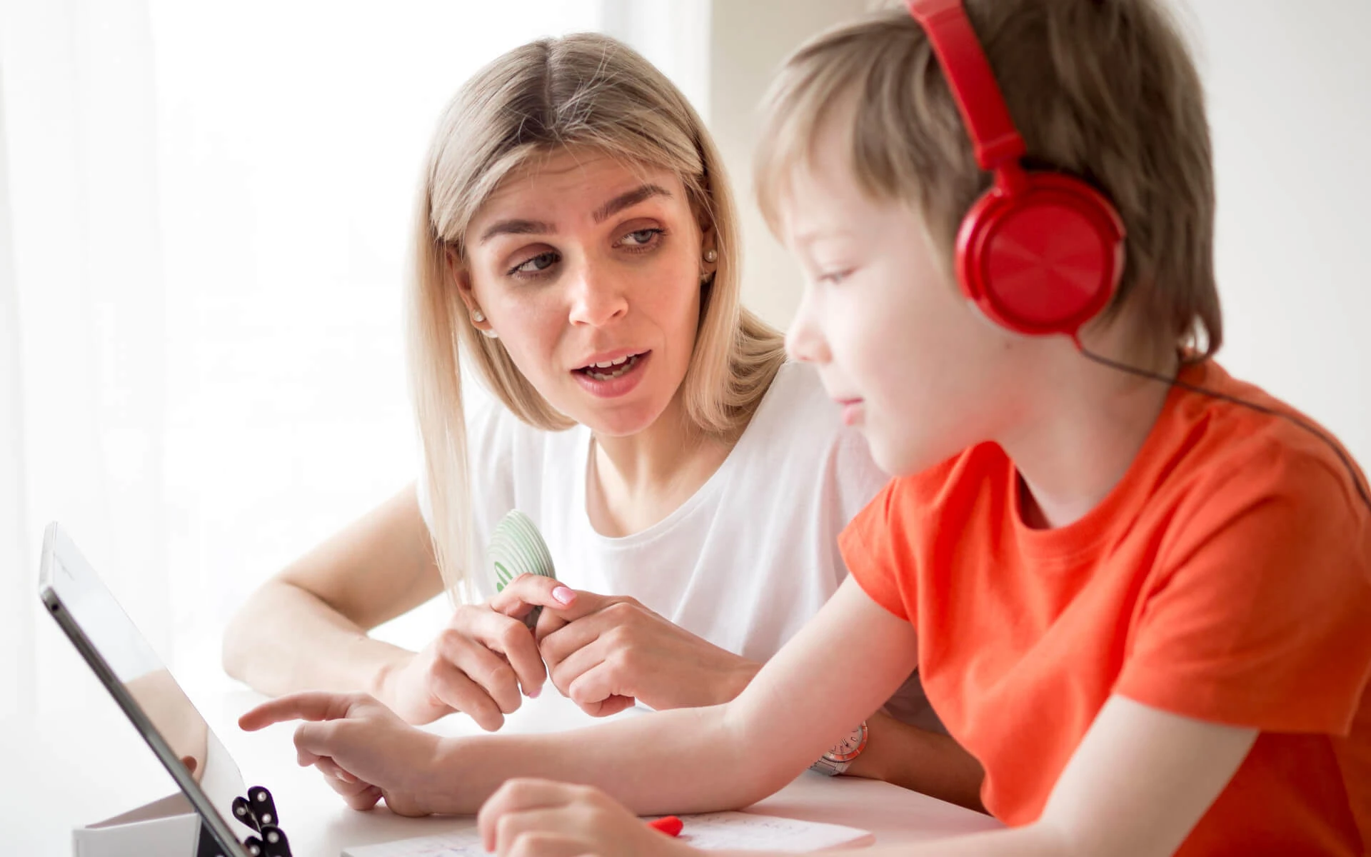 Eine Schulbegleiterin sitzt neben einem Jungen, der gerade rote Kopfhörer auf hat.