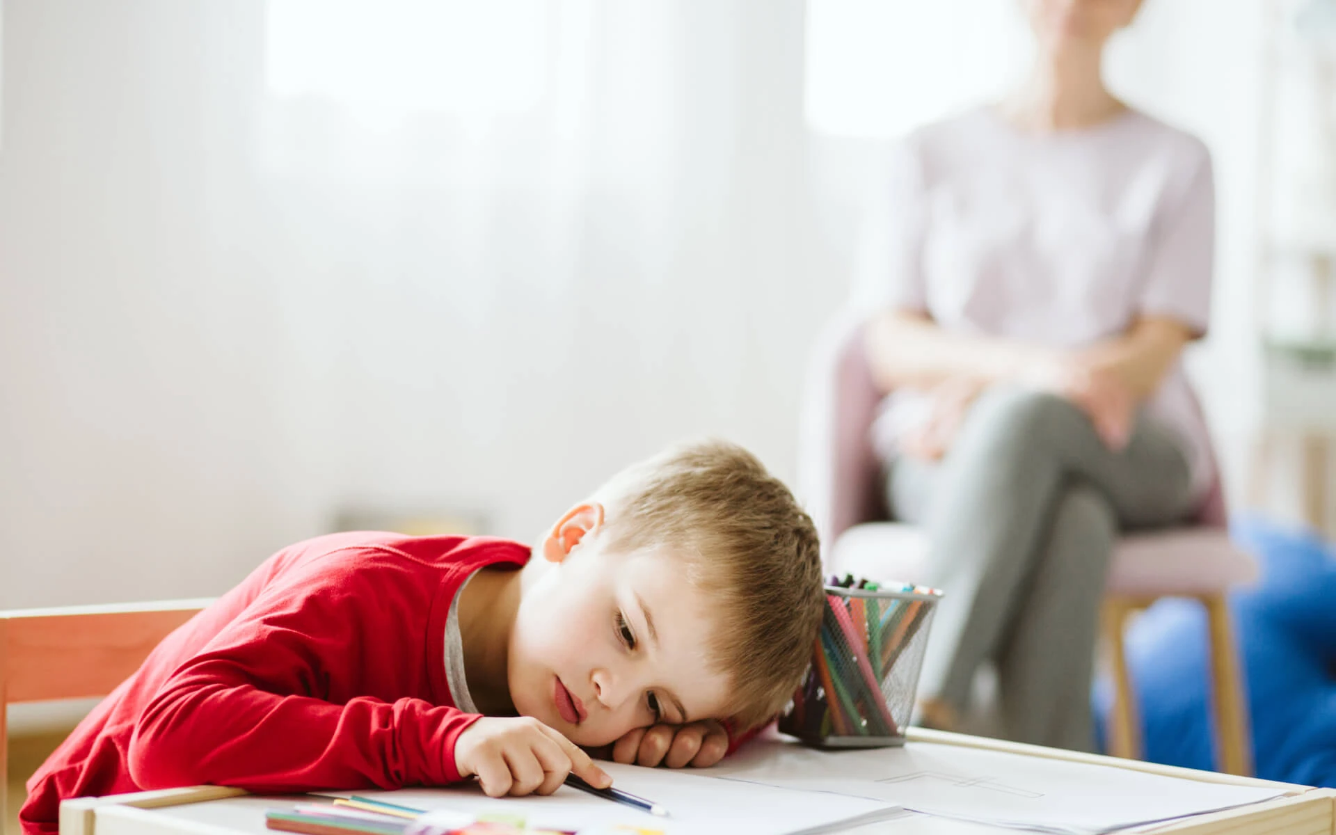 Ein Kind liegt mit seinem Kopf auf seinem Heft und hat einen Stift in der Hand. Im Hintergrund sitzt eine Person und beobachtet es.
